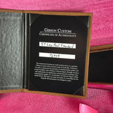 A Gibson Custom Certificate of Authenticity insert shown inside an open presentation folder on a pink textured fabric background. The black insert reads 'GIBSON CUSTOM CERTIFICATE OF AUTHENTICITY' and includes handwritten instrument identification and serial number 7893.