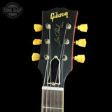 Close-up of a Gibson Les Paul HS-101 acoustic guitar headstock, featuring a black finish with yellow tuning pegs and gold Gibson branding, set against a solid black background.