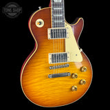 A close-up of a Gibson Les Paul Custom electric guitar body featuring a vibrant sunburst finish that transitions from bright amber-orange in the center to a dark reddish-brown edge. The guitar has a cream pickguard, gold hardware, and chrome hardware, shown against a solid black background with a Tone Shop watermark in the top left corner.