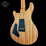A close-up of an electric guitar body showing a natural wood grain finish with black pickguard plates, photographed against a solid black background. A Tone Shop watermark is visible in the top left corner.