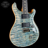 A close-up of an electric guitar body with a blue and silver flamed maple top, black pickups, and a black bridge, shown against a solid black background. A Tone Shop watermark is visible in the top-left corner.