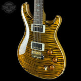 A close-up of an electric guitar body with a glossy brown and amber flame maple wood finish, featuring gold hardware, white pickguard, and a dark fretboard, shown against a solid black background.