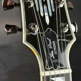 Close-up of a guitar headstock with six tuning pegs, strings, and a black pickguard featuring the text 'Les Paul SUPREME' and 'Gibson' in script.