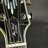 Close-up of a Les Paul Supreme electric guitar headstock, showing the black glossy finish, mother-of-pearl inlays, tuning pegs, and the Les Paul Supreme logo on the pickguard.