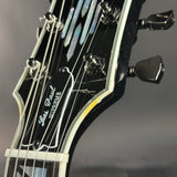 Close-up of the headstock of a Les Paul Supreme electric guitar, showing the black glossy finish, white binding, mother-of-pearl inlays, and tuning pegs against a dark gray background.