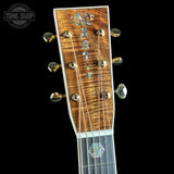 Close-up of a Hawaiian guitar headstock featuring a vibrant, curly figured top in natural brown wood, iridescent abalone shell inlays spelling 'HARIPUNA', and gold-toned tuning pegs against a solid black background.