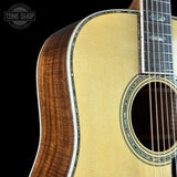 Close-up of an acoustic guitar body showing the natural wood side, soundboard, and part of the fretboard against a black background, with a Tone Shop watermark in the top left.