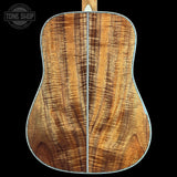 Close-up of the back of an acoustic guitar showing a richly figured wood grain top, with a white binding edge, set against a solid black background. A Tone Shop watermark is visible in the top left corner.