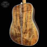 The back of an acoustic guitar made of figured wood, showing a rich grain pattern in warm brown and golden tones, set against a solid black background with a Tone Shop watermark in the top left corner.
