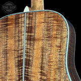 Close-up of the back of an acoustic guitar showing a rich, figured wood grain finish with a silver-colored metal binding edge against a black background.