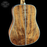 The back of an acoustic guitar with a rich, figured wood grain top, shown against a solid black background with a Tone Shop watermark in the upper left.