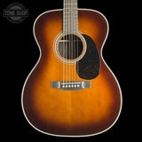 A close-up studio shot of an acoustic guitar body against a black background, showing a natural sunburst finish that fades from bright amber-orange at the edges to a darker brown center, with a black pickguard and white binding.