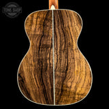 Close-up of the back of an acoustic guitar showing a rich, figured wood grain pattern in warm brown and golden tones, set against a solid black background with a Tone Shop watermark in the top left corner.