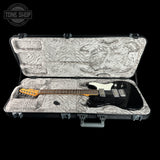 An electric guitar with a black body and white pickguard resting inside an open hard case with a textured gray plush lining. The guitar has an orange headstock.