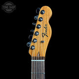 Close-up of a Fender electric guitar headstock with six black tuning pegs and a natural wood finish, shown against a solid black background.