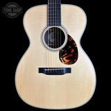An acoustic guitar with a natural wood finish and a tortoiseshell pickguard, shown against a solid black background with a Tone Shop logo in the top-left corner.