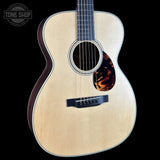 A close-up studio shot of an acoustic guitar body against a black background, showing a natural wood grain top, dark sides, a black bridge, and a tortoiseshell-style pickguard. A Tone Shop watermark is visible in the upper left corner.