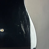 Close-up of a glossy black guitar body showing visible scratches and scuffs on the finish, with a small gold-toned hardware detail on the left against a neutral gray background.