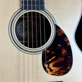 Close-up of an acoustic guitar body showing the round soundhole with decorative rosette, six strings, a dark fingerboard, and a tortoiseshell-style pickguard on a light natural wood finish.