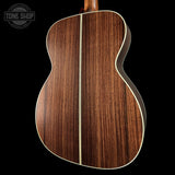 Close-up of the back of an acoustic guitar showing a rich dark brown wood grain finish with a light cream binding, set against a solid black background. A Tone Shop watermark is visible in the top-left corner.