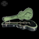 An acoustic guitar with a glossy black finish resting inside an open black hard case with a green velvet interior, shown against a solid black background. A Tone Shop logo is visible in the top-left corner.
