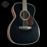 A close-up of a dark acoustic guitar body with a glossy black finish, white binding, a brown pickguard, and a silver bridge, shown against a black background with a Tone Shop watermark in the top left.