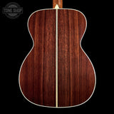 The back of an acoustic guitar made of rich reddish-brown wood with a light cream binding, shown against a solid black background. A Tone Shop watermark is visible in the top-left corner.