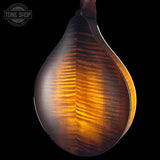 Close-up of a teardrop-shaped acoustic guitar body showing a rich, figured wood grain pattern in warm amber and brown tones against a solid black background. A Tone Shop watermark is visible in the top-left corner.