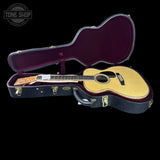 An acoustic guitar with a natural wood body and dark neck, displayed inside an open black hard case with a purple interior, set against a solid black background. A Tone Shop watermark is visible in the top-left corner.