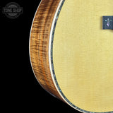 Close-up of the lower portion of an acoustic guitar, showing the natural wood side edge, the light gold-toned top, and part of the black f-hole cover on a black background. A Tone Shop watermark is visible in the upper left.