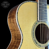 Close-up of an acoustic guitar body showing the natural wood grain back, soundhole rosette, and part of the dark fretboard against a black background. A Tone Shop watermark is visible in the top-left corner.