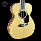 An acoustic guitar with a natural wood finish and a dark wood sides, shown against a solid black background. A circular 'TONE SHOP' watermark is visible in the top-left corner.