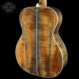 Close-up of the back of an acoustic guitar showing a rich, figured wood grain finish in warm brown and golden tones, with a white binding edge, set against a solid black background.