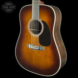 A classic acoustic guitar with a sunburst finish, featuring a dark brown center, lighter amber edges, and a black pickguard, shown against a solid black background with a Tone Shop watermark in the top left corner.