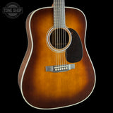 A close-up studio shot of an acoustic guitar body against a black background, showing a natural sunburst finish that fades from bright amber-orange in the center to darker brown at the edges. The guitar features a black pickguard, a silver-toned bridge, and white binding around the body and neck.