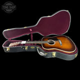 An acoustic guitar with a sunburst finish displayed inside an open hard case with a burgundy velvet interior, set against a solid black background. A Tone Shop watermark is visible in the top-left corner.