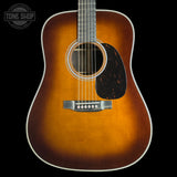 A classic acoustic guitar with a natural sunburst finish, featuring a dark black pickguard and a white binding around the body, shown against a solid black background with a Tone Shop logo in the top left corner.