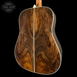 The back of an acoustic guitar featuring a rich, symmetrical wood grain pattern in warm brown and golden tones, with a white binding edge, shown against a solid black background.