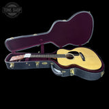 An acoustic guitar with a natural wood top and dark neck, displayed inside an open black hard case with a burgundy interior, set against a solid black background. A Tone Shop watermark is visible in the top-left corner.