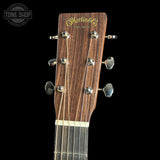 Close-up of the headstock of a Martin & Co acoustic guitar against a solid black background, showing the brand name in gold script and six silver tuning pegs.