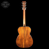 A wooden acoustic guitar shown from the back, featuring a rich warm brown wood grain finish and six tuning pegs on the headstock, set against a solid black background with a Tone Shop watermark in the top left corner.