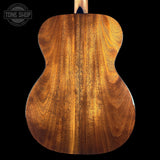 Close-up of the back of an acoustic guitar showing a rich, warm brown wood grain finish, set against a solid black background with a Tone Shop watermark in the top left corner.