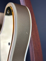 Close-up detail of an electric guitar body showing a metallic champagne gold finish, a white edge trim, and a dark blue background.