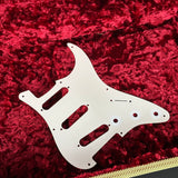 A white plastic guitar pickguard with pre-drilled holes, displayed on a plush red velvet surface.