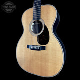 Acoustic guitar with a black pickguard on a black background, featuring the Tone Shop logo.