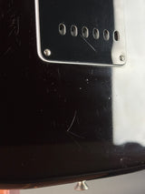 Close-up of the glossy black body of an electric guitar, showing a silver metal plate with five oval cutouts and visible surface scratches.