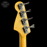 Close-up of the headstock of a bass guitar with a natural maple wood finish and four silver metal tuning pegs, shown against a solid black background. A Tone Shop watermark is visible in the top left corner.