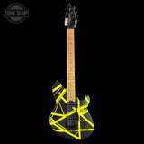 A black electric guitar with a bold yellow geometric tape-like graphic pattern on the body, shown against a solid black background. The headstock is black with silver tuning pegs, and the neck is a natural maple wood finish.