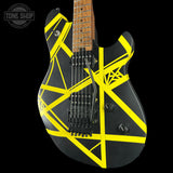 A black electric guitar with bold yellow geometric tape-like graphics across the body, shown against a black background. The neck appears to be a natural wood tone, and a Tone Shop watermark is visible in the upper left corner.
