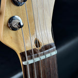 Close-up detail of a guitar headstock and neck, showing the natural wood body, dark fretboard, silver tuning pegs, and strings against a dark background.